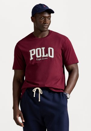 Mann iført en burgunder Polo Ralph Lauren T-skjorte, marineblå joggebukser, marineblå caps og klokke, står mot en klar lys bakgrunn.