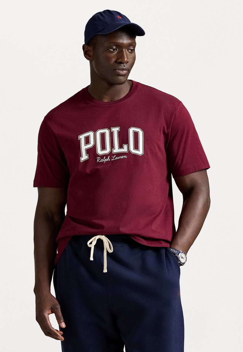 Férfi burgundi Polo Ralph Lauren pólót, tengerkék húzózsinóros nadrágot, tengerkék sapkát és órát visel, egy sima világos háttér előtt áll.