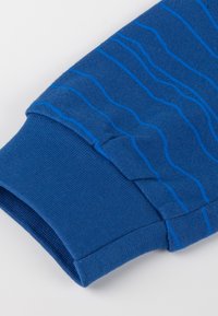 Manica di un maglione blu di cotone con polsino a coste testurizzato e design a onde in un blu più chiaro. Finitura del tessuto liscia.