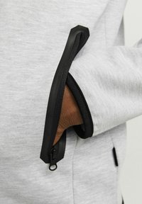 Felpa grigia con una tasca con zip nera e un accento in gomma, che mette in evidenza un tessuto testurizzato e un dettaglio della zip in contrasto.