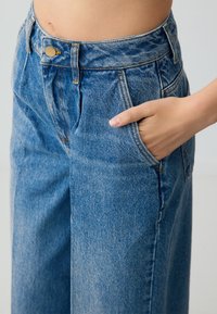 Jean large taille haute en denim bleu clair, avec un bouton doré et des poches frontales visibles. Présente une texture légèrement délavée.