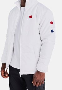 Man som bär en vit zip-up jacka med små röda, vita och blå blommönster på bröstet och vänster ärm, kombinerad med svarta byxor.