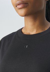 Close-up van het onderste deel van het gezicht en de nek van een persoon die een zwart shirt met ronde hals draagt, met een klein geborduurd Champion-logo op de borst.