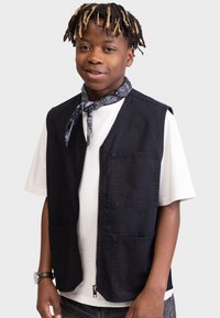Gilet senza maniche in denim nero con due tasche frontali, chiusura con zip e un tessuto strutturato. Indossato sopra una t-shirt bianca e accessorGuardato con una sciarpa a fantasia.