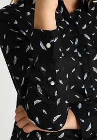 Chemise noire ornée d'un motif de plumes blanches, avec des poignets à boutons et des détails au col. Le tissu semble lisse avec un subtil éclat.