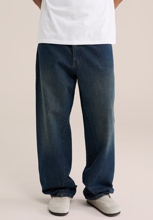 Personne portant un jean bleu foncé ample, une chemise blanche et des chaussures slip-on gris clair, debout sur un fond neutre.