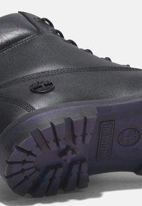 Botte en cuir noir avec semelle épaisse à crampons, logo arbre Timberland sur le côté et sur la semelle, coutures détaillées le long de la partie supérieure de la chaussure.