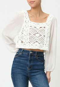 Top court avec un design en dentelle blanche au crochet présentant des motifs géométriques, associé à un jean taille haute bleu clair et à des manches longues transparentes.