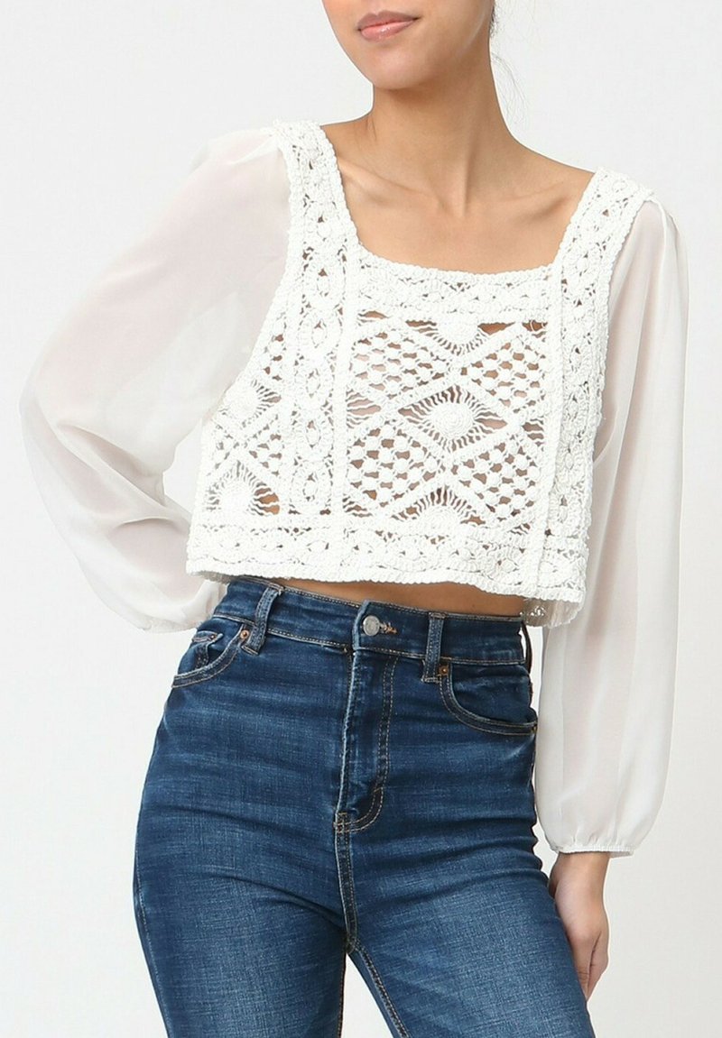 Top court avec un design en dentelle blanche au crochet présentant des motifs géométriques, associé à un jean taille haute bleu clair et à des manches longues transparentes.