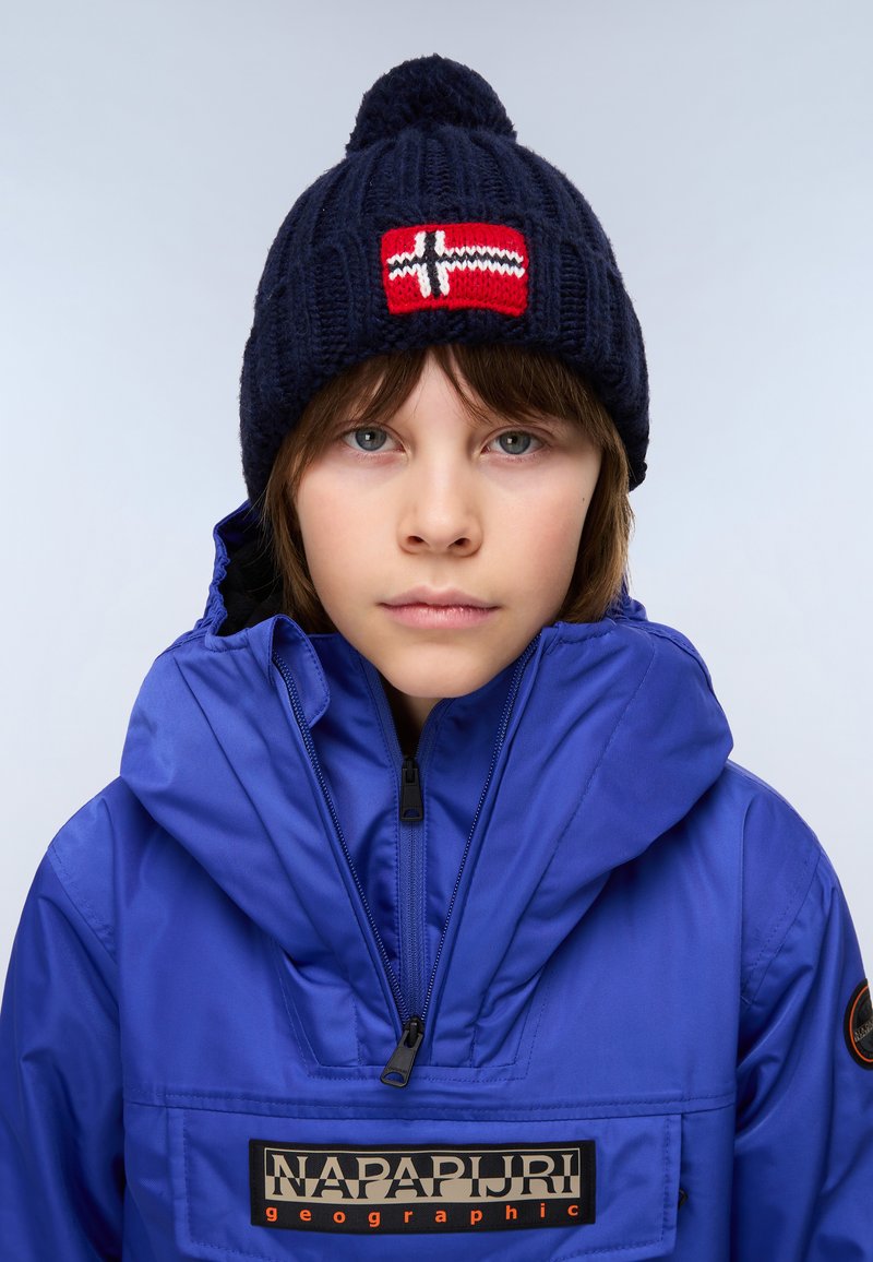 Child wearing a blue Napapijri jacket and a navy knit hat with a red and white flag patch, facing forward against a plain background.