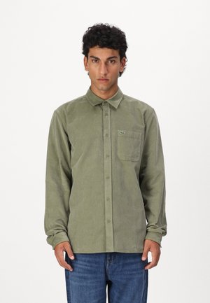 Camisa de pana verde con cuello abotonado, bolsillo delantero y logo bordado. Mangas largas y dobladillo recto. Llevada con jeans azules.