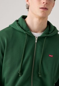 Felpa verde con zip e coulisse; realizzata in tessuto morbido; presenta un piccolo logo patch rosso di Levi's sul petto; vestibilità normale.