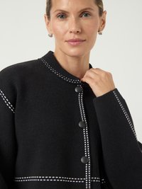 Γυναίκα που φοράει μαύρη καπιτονέ ζακέτα με λευκές ραφές, κρατώντας τον γιακά κοντά στον λαιμό της, σε ουδέτερο φόντο.