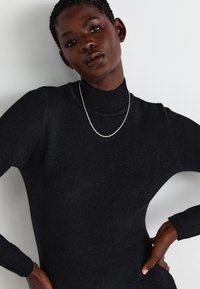 Fekete, testhez simuló garbó pulóver bordás mintázattal és hosszú ujjakkal, ezüstlánc nyaklánccal párosítva. Egyszerű dizájn, minimális kiegészítők.