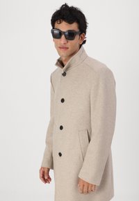 Abrigo de mezcla de lana beige con un cuello alto, cinco botones negros, bolsillos laterales y un diseño ligeramente estructurado. Lo lleva puesto con gafas de sol negras.