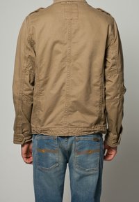 Bawełniana kurtka khaki z pagonami na ramionach, o prostym kroju i długich rękawach. Połączona z niebieskimi dżinsami z kontrastowymi przeszyciami.