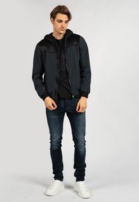 Giubbotto bomber nero con zip e dettagli in rete, abbinato a una t-shirt grafica nera, jeans denim scuri e sneakers bianche.