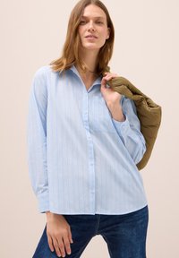 Camicia azzurra a righe con bottoni, caratterizzata da maniche lunghe, tasca sul petto e una vestibilità rilassata; indossata con un giubbotto trapuntato verde sulle spalle.