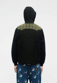 Μαύρο φούτερ με κουκούλα, με πράσινη καρό λεπτομέρεια στους ώμους, υφασμάτινη υφή, συνδυασμένο με μπλε τζιν που έχουν λευκά σχέδια yin-yang.