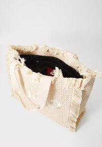 Woven tote bag in light beige with fringed edges, featuring a black interior and textured exterior, includes handles for easy carrying.