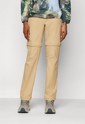 Pantaloni beige leggeri da outdoor con gambe rimovibili per versatilità, realizzati in tessuto liscio. Indossati con scarpe da trekking robuste.