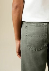 Olivzaļas kokvilnas bikses ar vienu aizmugures kabatu. Audumam ir smalka tekstūra un redzama šuvju apdare ap kabatas malu.