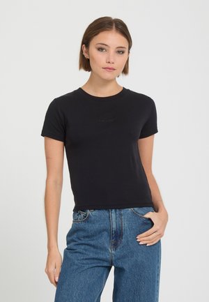 Jeune femme aux cheveux courts bruns portant un t-shirt noir uni à manches courtes et un jean bleu taille haute, une main dans la poche.