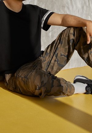 Osoba siedząca na żółtej podłodze w kamuflażowych spodniach, czarnym T-shircie w białe paski, białych skarpetkach i czarnych trampkach.