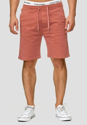 Homme portant un short rouge délavé à cordon, un sous-vêtement blanc avec ceinture Calvin Klein et des baskets blanches, debout devant un fond uni.