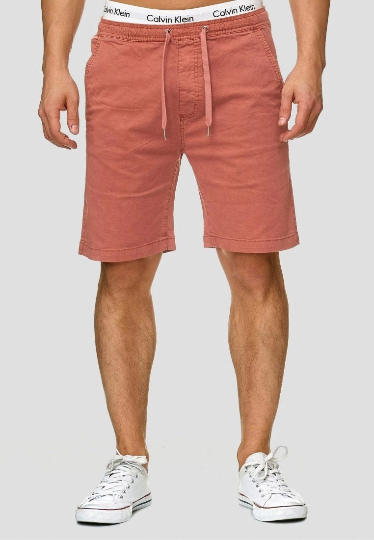 Homme portant un short rouge délavé à cordon, un sous-vêtement blanc avec ceinture Calvin Klein et des baskets blanches, debout devant un fond uni.