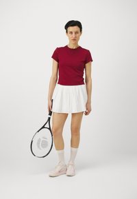 Punane lühikeste varrukatega pluus koos valge voltidega seelikuga. Käsikäes must tennisehaamer. Roosad tossud valgete ja heleroosade detailidega.