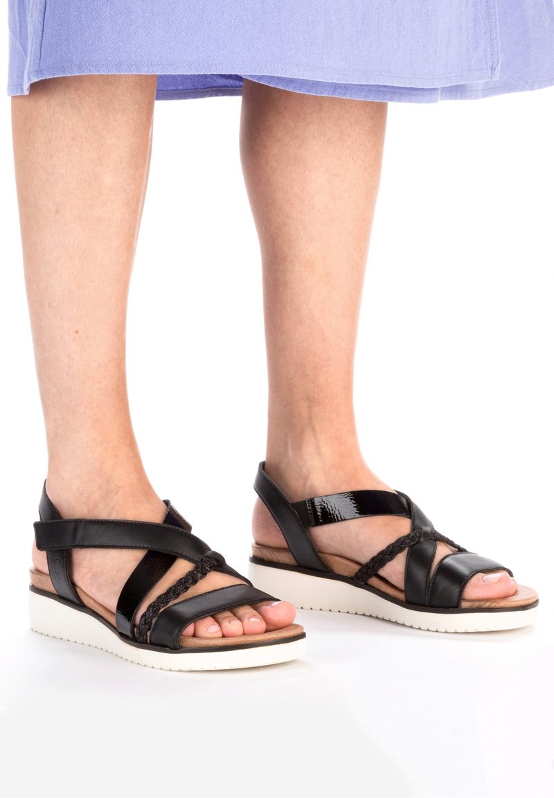 Personne portant des sandales compensées noires à brides avec des semelles blanches et une jupe violet clair, debout sur une surface blanche.