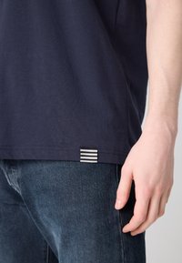 T-shirt en coton marine avec une coupe droite, featuring une étiquette rayée en bas. Associé à un jean en denim foncé. Texture minimale, design simple.