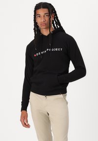 Zwarte hoodie met witte "DENIM PROJECT" tekst, voorzien van een voorkant kangoeroezak en verstelbare trekkoorden. Draagt bij beige broeken.