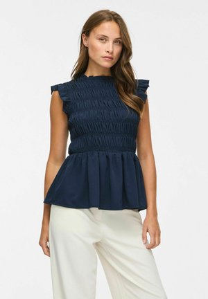 Femme portant un haut sans manches bleu marine froncé avec des épaules à volants et un pantalon blanc à jambes larges sur un fond uni.