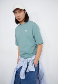 Tricou din bumbac verde deschis cu mâneci scurte și un mic logo alb. Șorturi din denim albastru cu o cămașă cu dungi albastre și albe legată în jurul taliei.