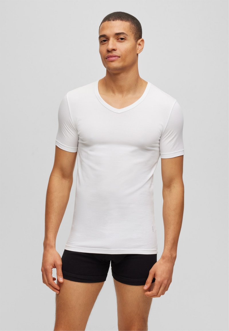 White short-sleeve V-neck t-shirt made of smooth cotton fabric, fitted design, paired with black shorts, model stands against a neutral background.