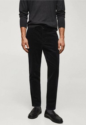 Mann trägt schwarze Cordhose, schwarze Loafers, dunkelgraues Langarmhemd, steht vor einem schlichten hellen Hintergrund.