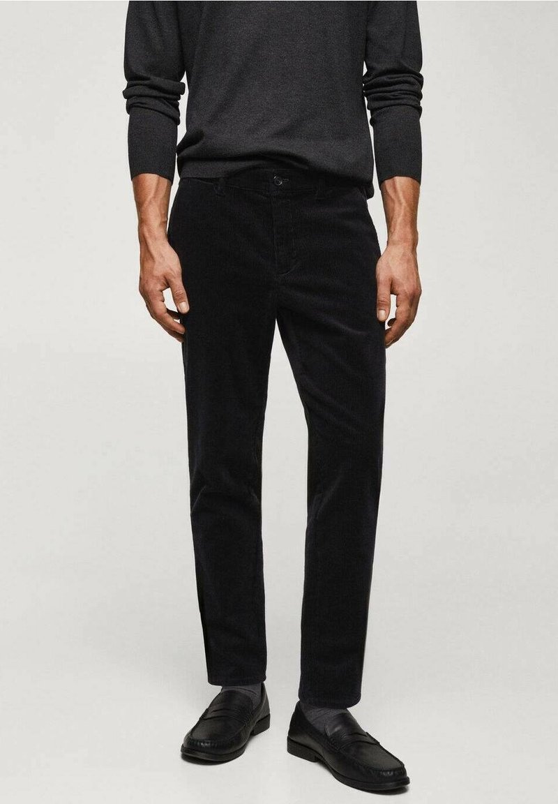 Homme portant un pantalon en velours côtelé noir, des mocassins noirs et une chemise à manches longues gris foncé, debout devant un fond clair uni.