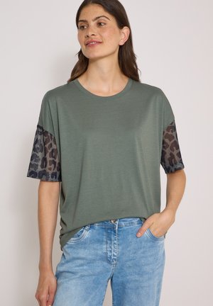 Femme aux longs cheveux bruns portant un t-shirt vert à manches courtes avec manches en maille à imprimé léopard et un jean bleu clair, une main dans la poche.