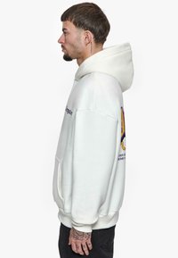 Hoodie blanc avec une poche kangourou, grande capuche et un imprimé graphique présentant du texte violet et un design coloré sur la manche gauche.