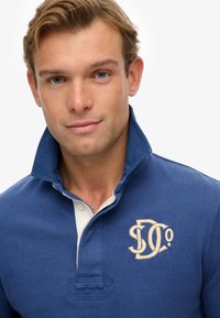 Blue polo shirt with a white collar, featuring a textured fabric and embroidered logo in beige. The collar is raised, emphasizing the design.