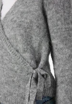 Krupan kadar sivo pleteni pulover z vezanjem sa strane preko plavih traperica, koji prikazuje teksturu i detalje tkanine.