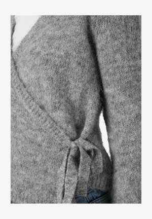 Krupan kadar sivo pleteni pulover z vezanjem sa strane preko plavih traperica, koji prikazuje teksturu i detalje tkanine.