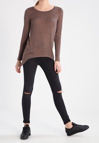 Braunes langärmliges Oberteil mit Rundhalsausschnitt, kombiniert mit schwarzen engen Jeans mit Knieflicken und schwarzen Sportschuhen. Glatte Textur.