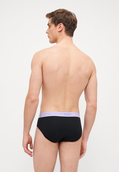 Slip in cotone nero con una vita elastica lilla decorata con il marchio "EMPORIO ARMANI", mostrato di spalle su un modello maschile.