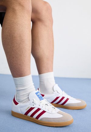 Beine mit weißen Socken und Adidas Samba Sneakers mit weißen, roten Streifen und Gummisohlen auf einem hellblauen Boden.