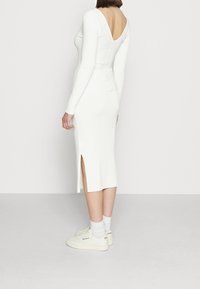 Bílá pruhovaná midi šaty s dlouhými rukávy, hlubokým výstřihem na zádech a bočními rozparky, doplněná o bílé tenisky a kotníkové ponožky.