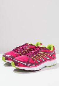 Ett par rosa och lila Salomon löparskor med limegröna accenter och vita sulor på en vit yta mot en grå bakgrund.