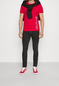 Červené bavlněné tričko s bílým logem "HILFIGER" na levé straně, v kombinaci s černými džíny a červenými teniskami s bílou podešví. Černý svetr přehozený přes ramena.